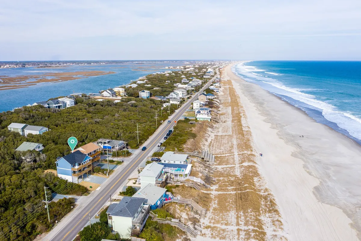 A tranquil stretch of beachfront with gently rolling ocean waves, sandy dunes, and a coastal road lined with beachfront properties. The scene is bathed in peaceful early morning light, evoking a sense of calm and openness.