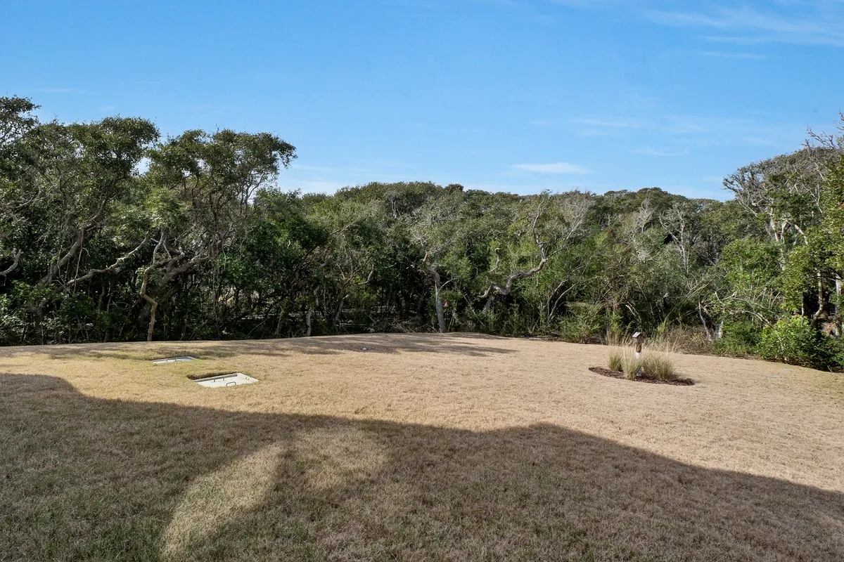 A spacious exterior lawn surrounded by mature trees and a clear blue sky overhead. The setting is serene and inviting, ideal for relaxing outdoors, watching sunsets, or enjoying nature walks with loved ones.
