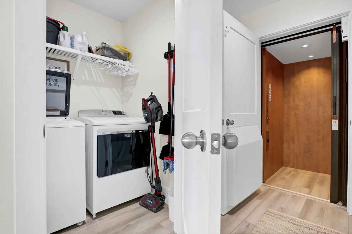 A practical, well-lit laundry area featuring a modern washing machine and dryer, with an elevator door conveniently located nearby. Cleaning supplies are neatly organized, emphasizing cleanliness and ease of use. The setting captures both essential laundry facilities and the added accessibility of the elevator.