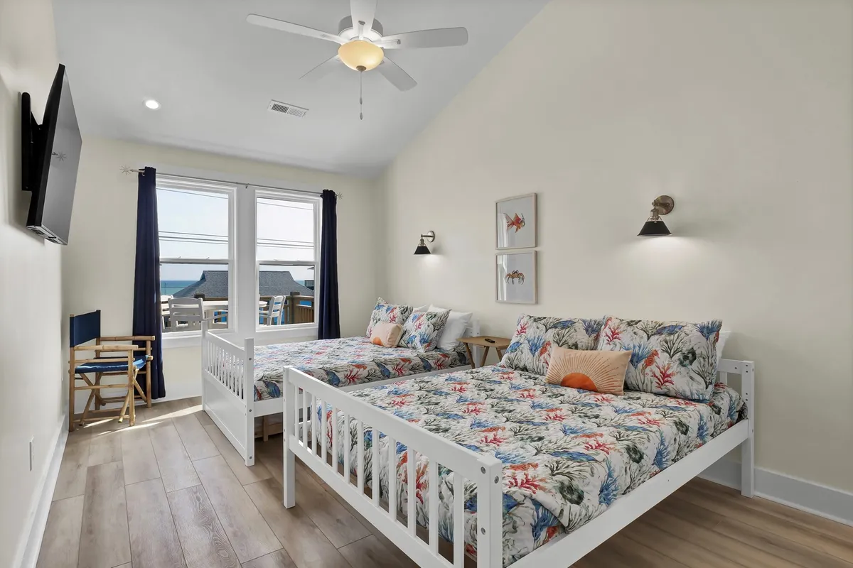 A cheerful bedroom with two full beds dressed in floral bedding, large windows offering ocean views, a wall-mounted TV, and inviting decor.