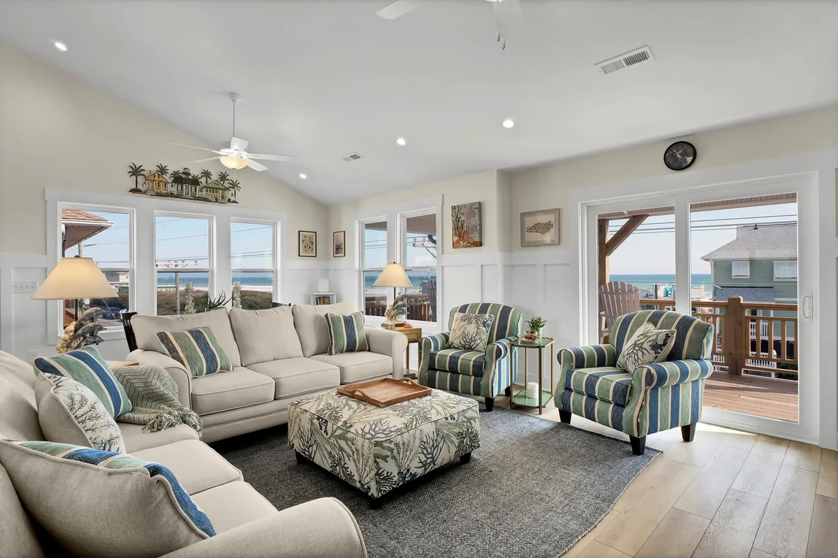 A spacious living room with a large sectional sofa, striped armchairs, and a decorative coffee table sits adjacent to expansive windows that showcase ocean views. The furnishings invite relaxation and socializing while keeping the beach scenery in constant view.