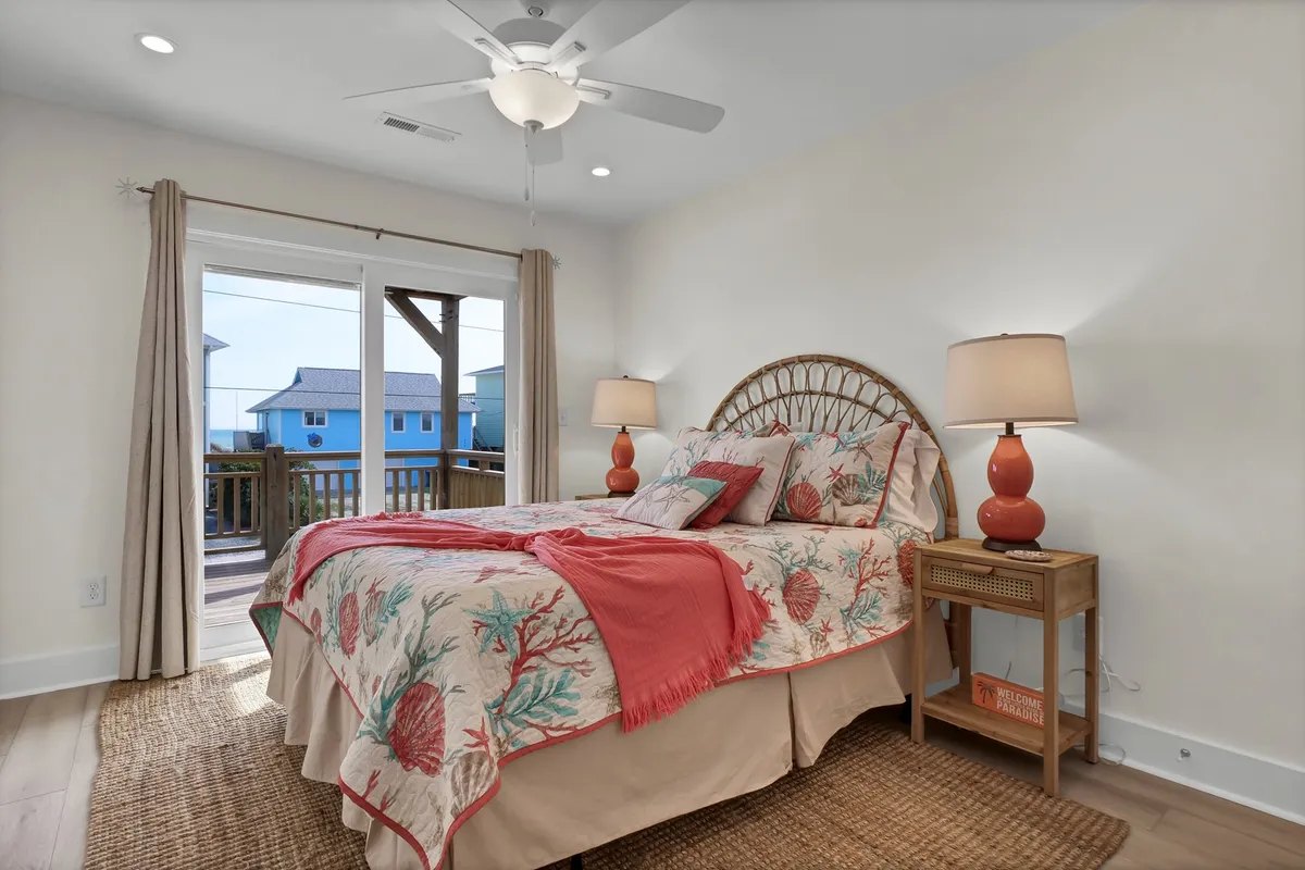 A tranquil bedroom with coral-patterned bedding, bedside lamps, and a view through glass doors that open directly to the deck, hinting at ocean vistas and gentle sea breezes.