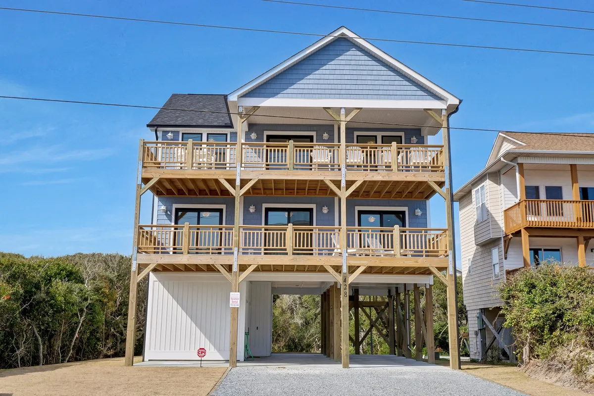 A cheerful three-story beach house with a wooden balcony and spacious deck, large windows, and a classic coastal exterior. The inviting atmosphere suggests a place for family gatherings, barbecues, and enjoying views after a day outdoors.