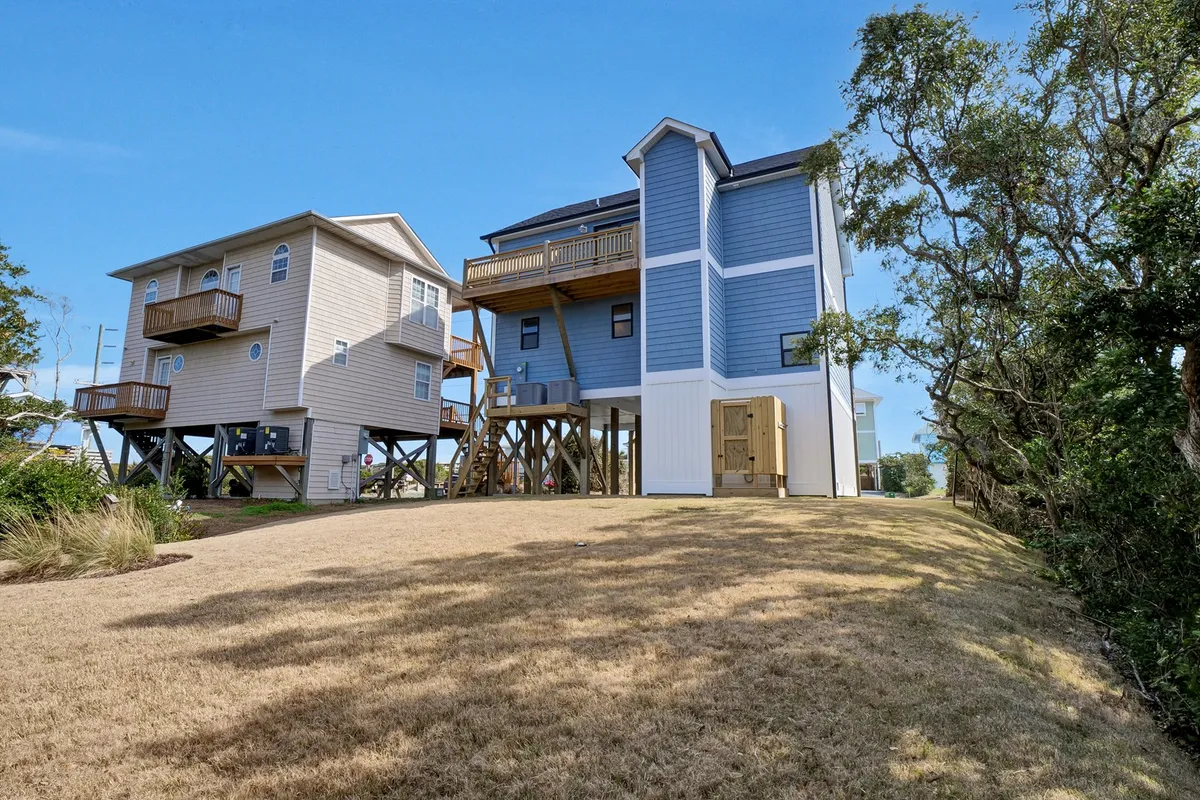 The image presents a pair of two-story houses with expansive wooden decks surrounded by greenery under a clear sky. It evokes a welcoming setting for outdoor barbeques, relaxation, and sunset or stargazing from the upper deck.